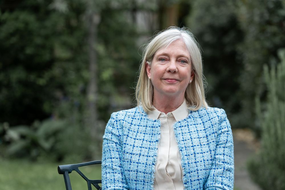A person wearing a light blue checkered blazer and white blouse sits outdoors against a green background.