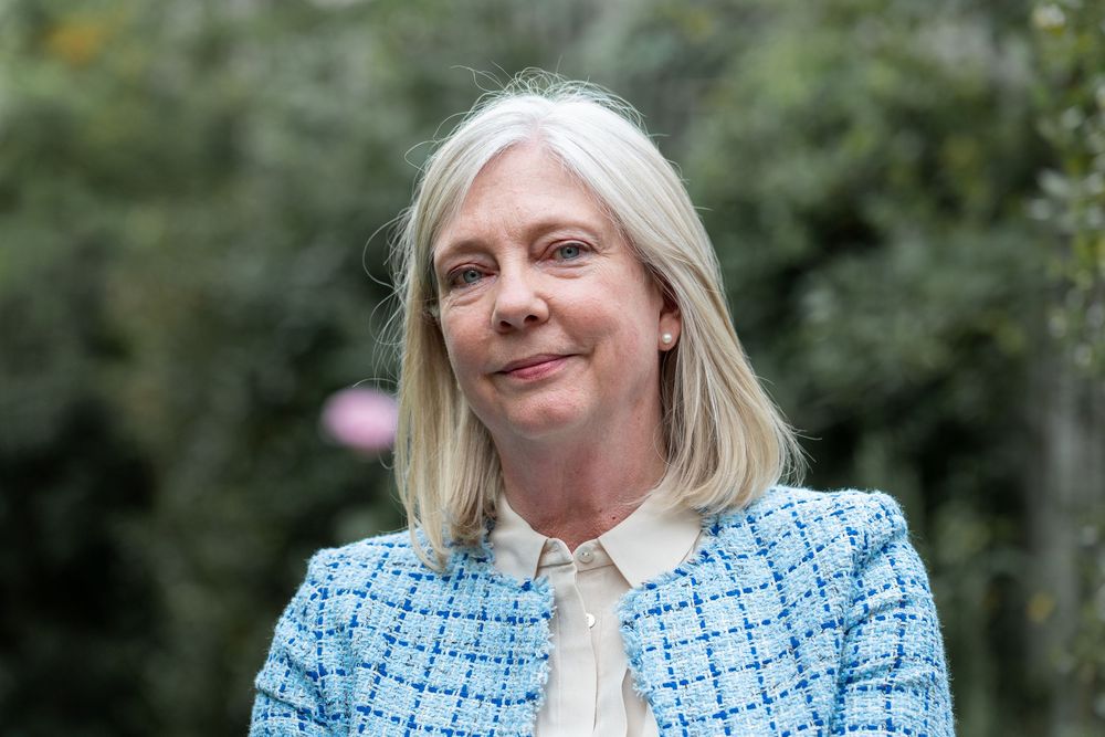A person in a blue tweed jacket poses outdoors against a natural green background.