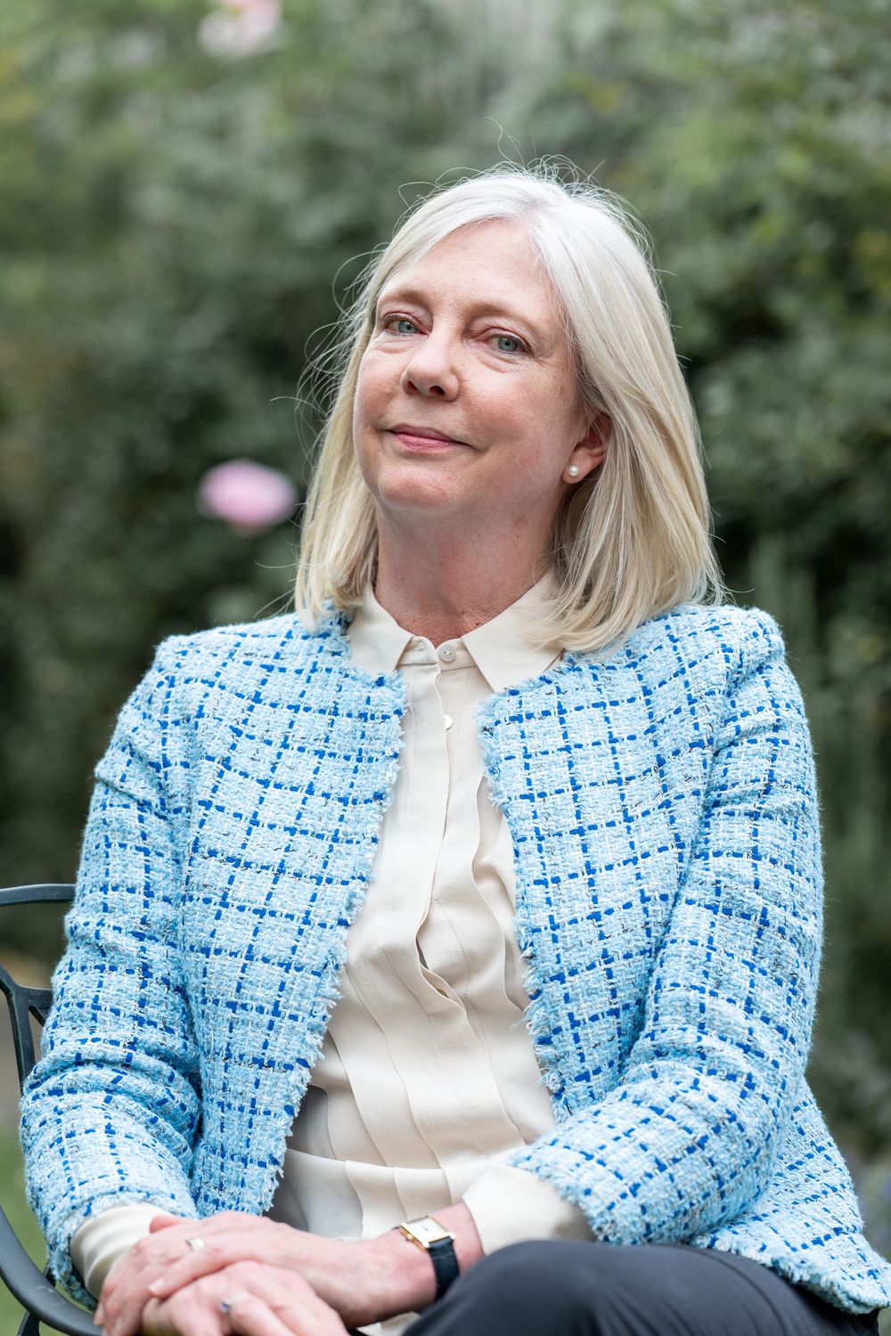 A person wearing a light blue tweed jacket and cream blouse sits outdoors against a garden background.