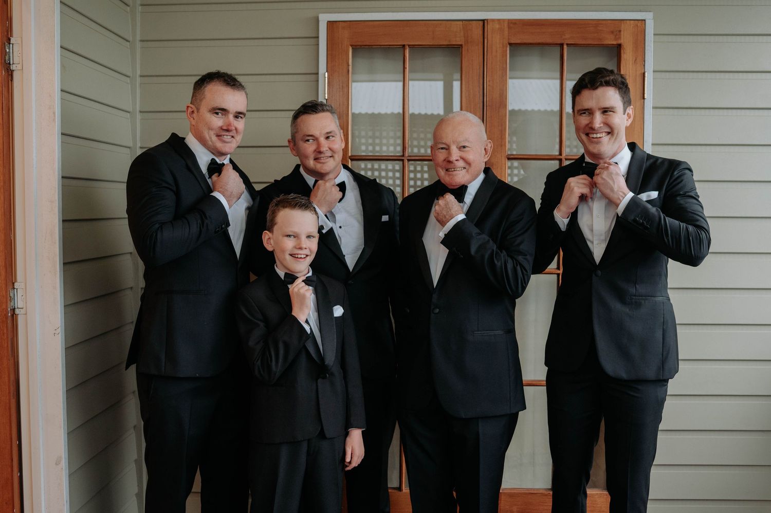Multigenerational group in formal black suits standing against beige wall while adjusting their bow ties.