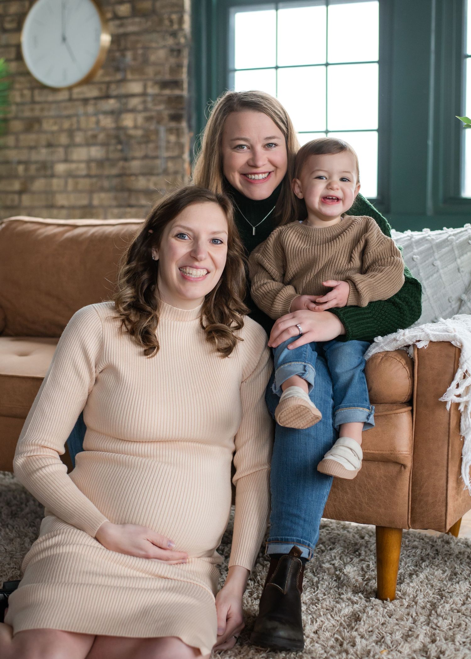 Three people sit together on a brown leather couch in a cozy living room with exposed brick walls and natural lighting.