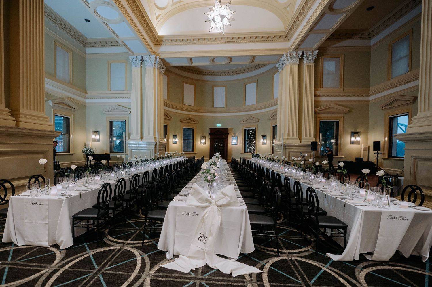 Elegant banquet hall with long tables arranged in two rows, featuring white linens and crystal chandeliers in a grand setting.