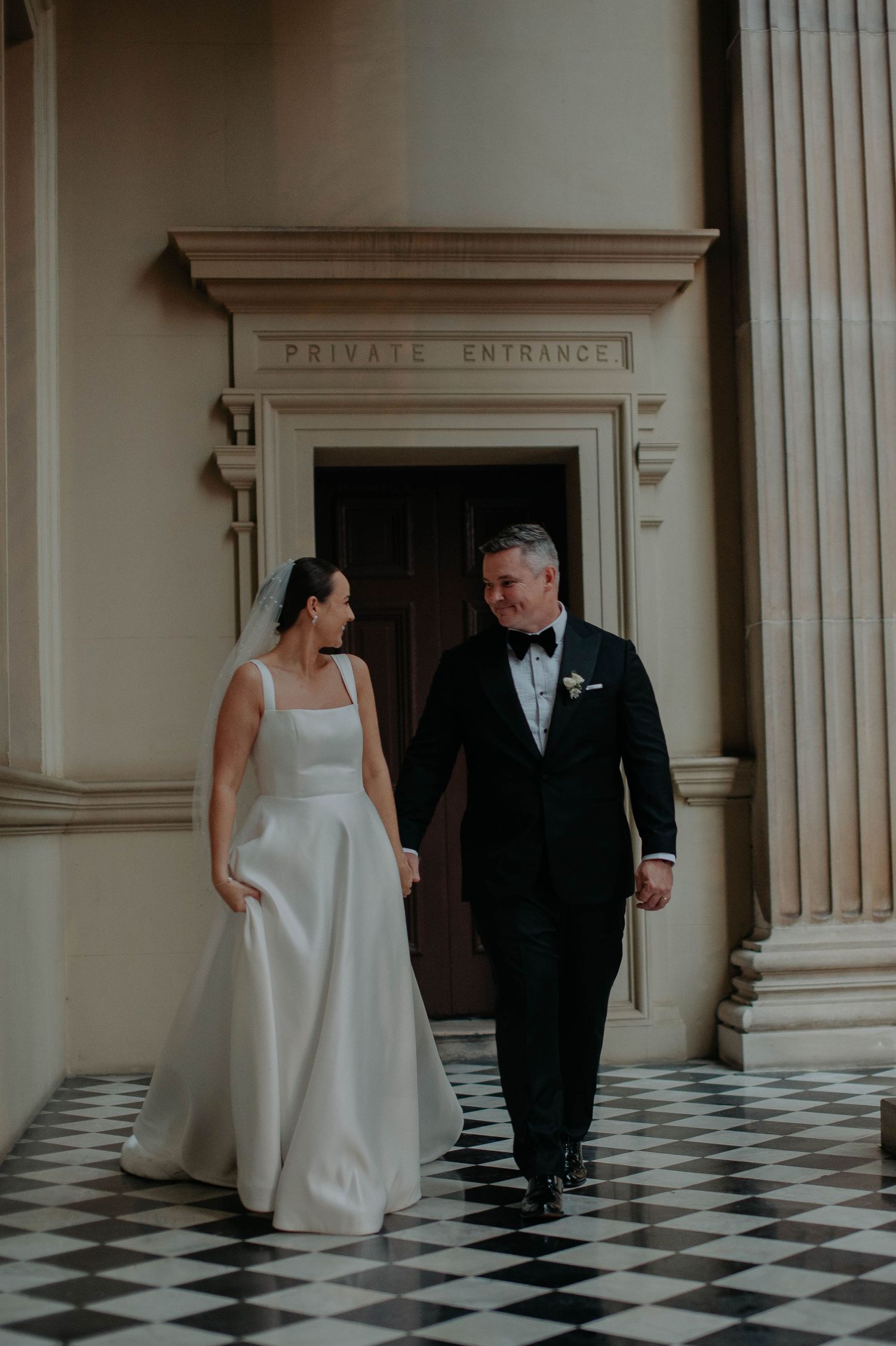 A couple in wedding attire share a romantic moment in front of a private entrance with checkered floor tiles.