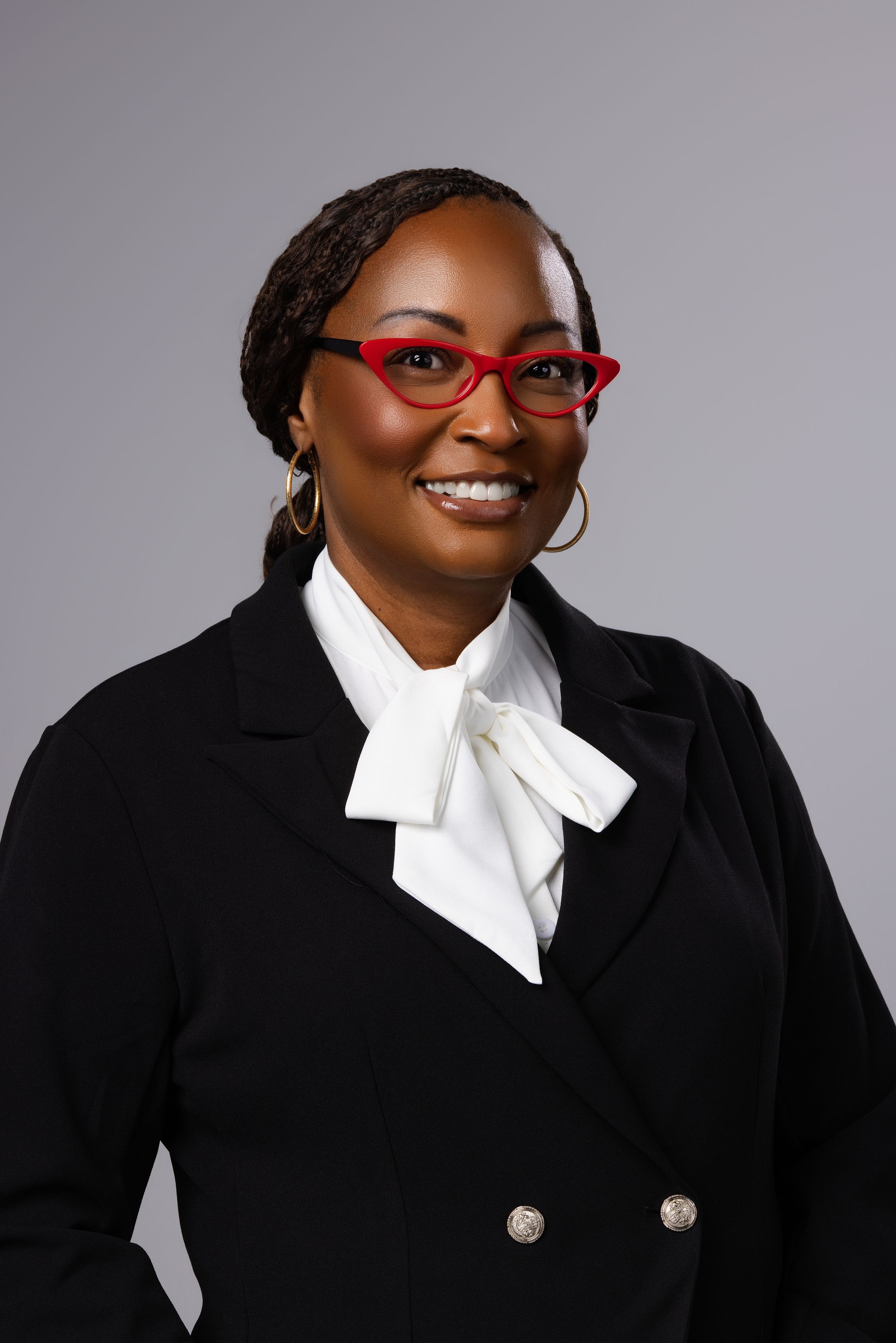 Professional woman in black blazer and red glasses smiling confidently against gray background.