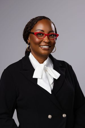 Professional woman in black blazer and red glasses smiling confidently against gray background.