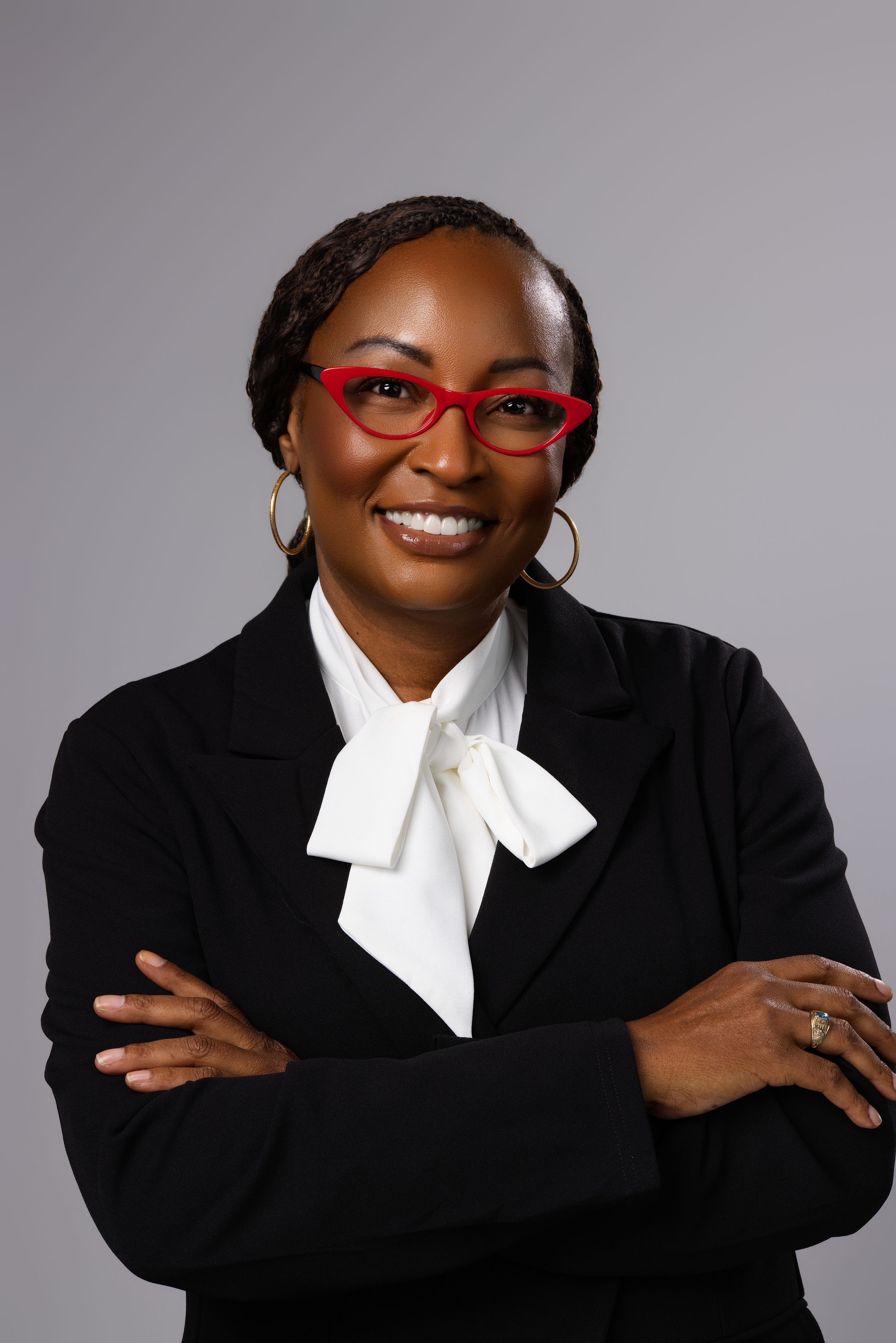 Professional woman in black blazer, red cat-eye glasses, and white bow blouse, smiling with arms crossed.