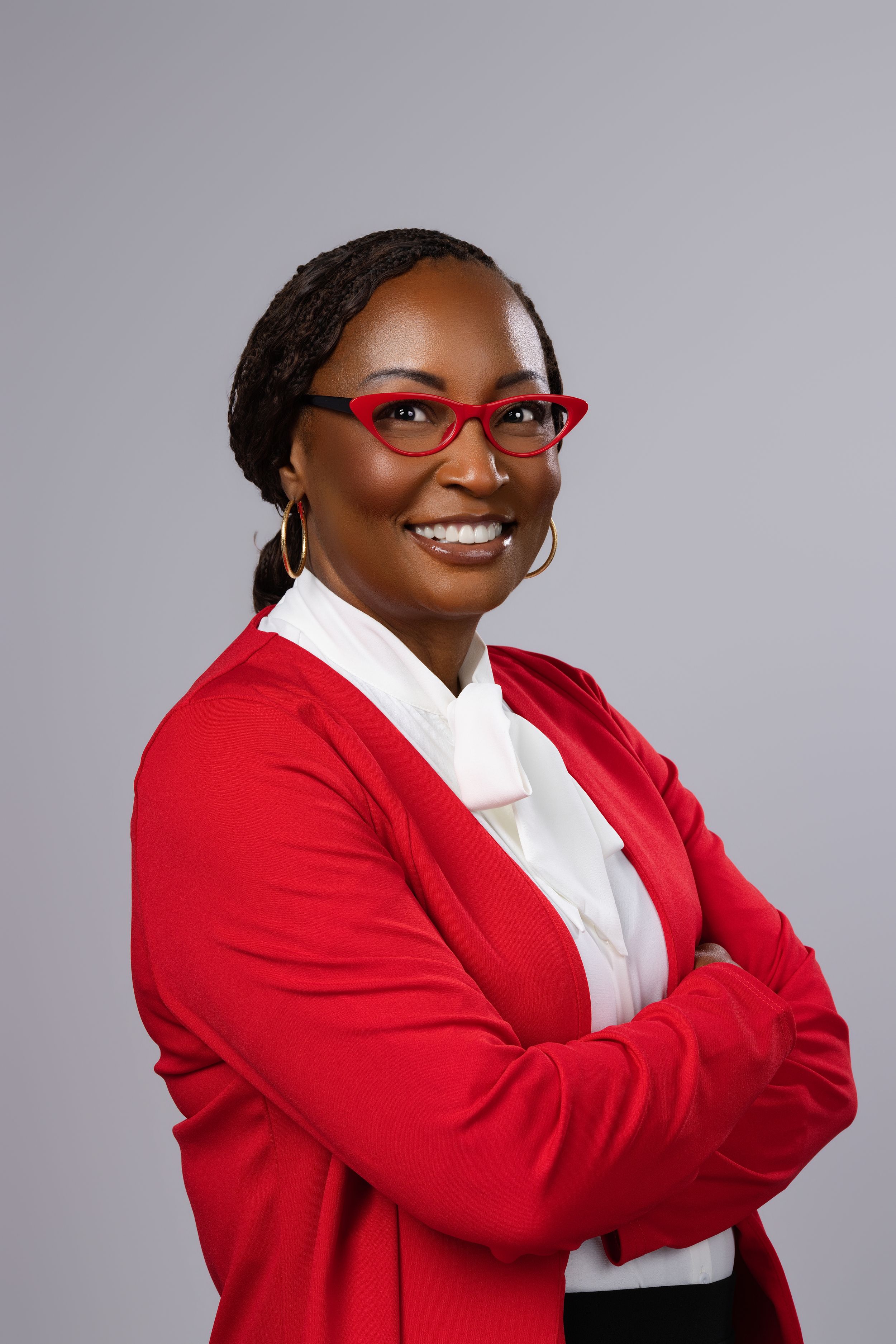 Professional woman in red blazer and glasses smiling with arms crossed against gray background.