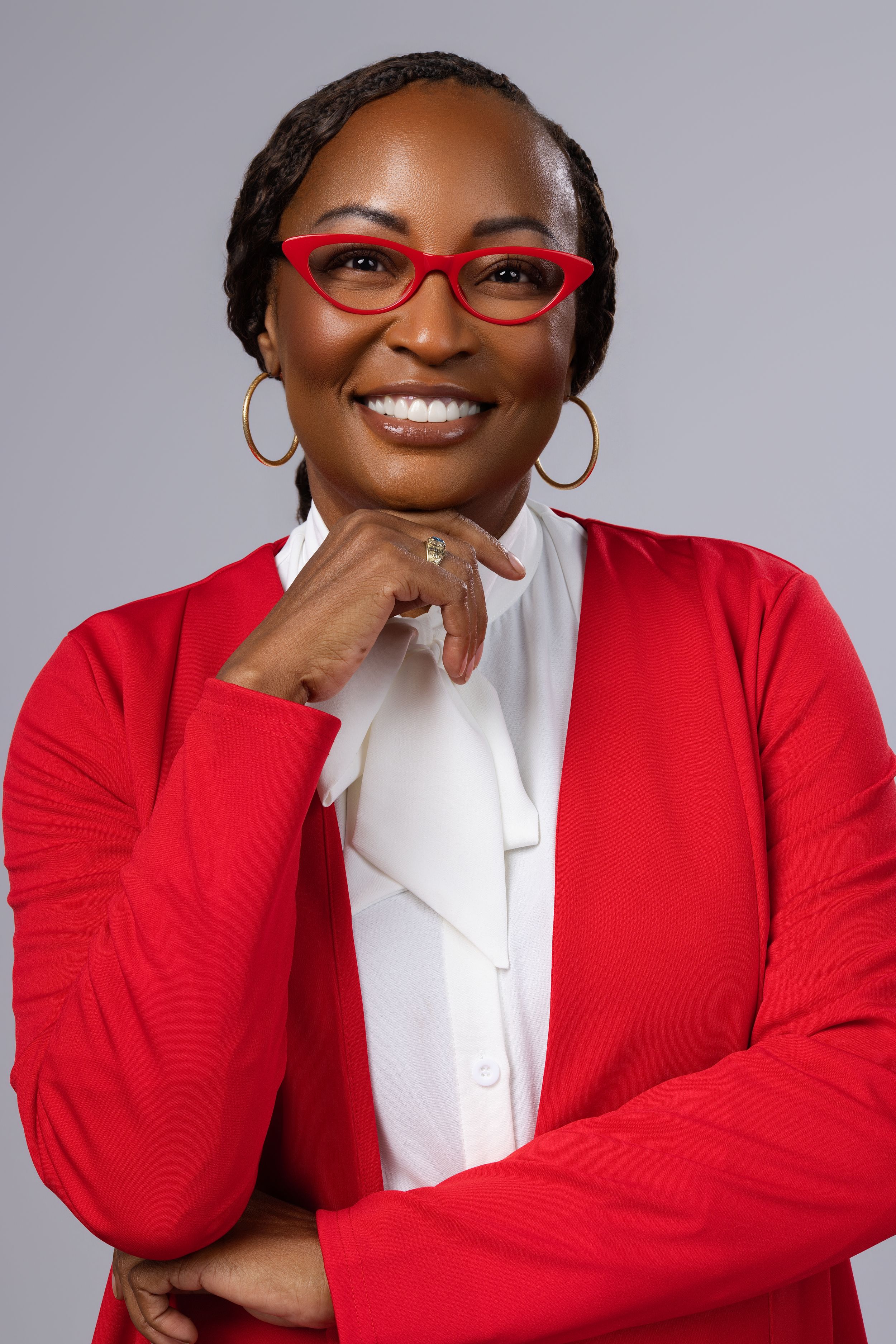 Smiling woman in red blazer and glasses with hand on chin poses confidently against gray background.