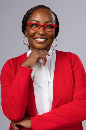 Smiling woman in red blazer and glasses with hand on chin poses confidently against gray background.