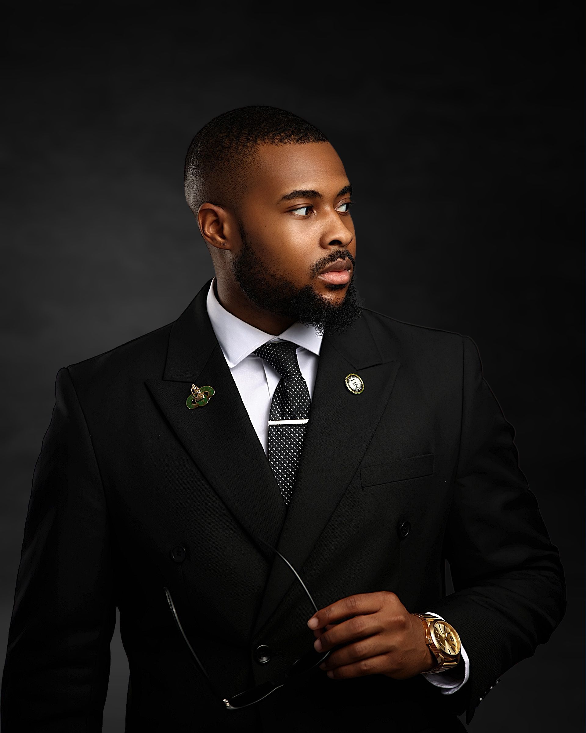 Confident man in black suit and tie with gold watch posing against dark background.