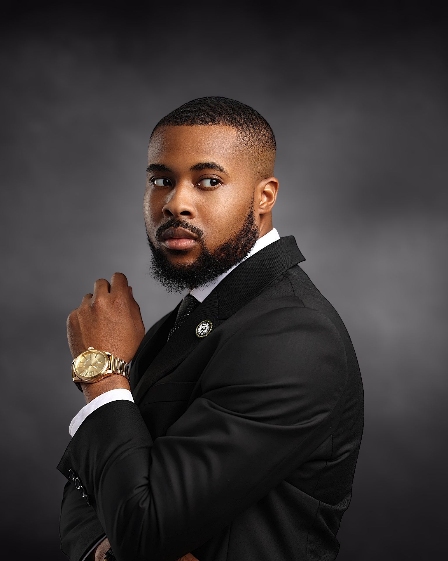 A well-dressed Black man in a black suit and gold watch poses confidently against a dark gray studio background.