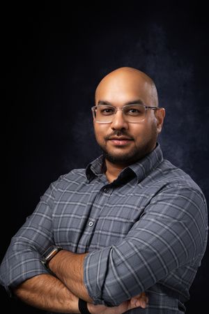 Bald man with glasses and crossed arms wearing a gray plaid shirt, posing confidently against a dark background.