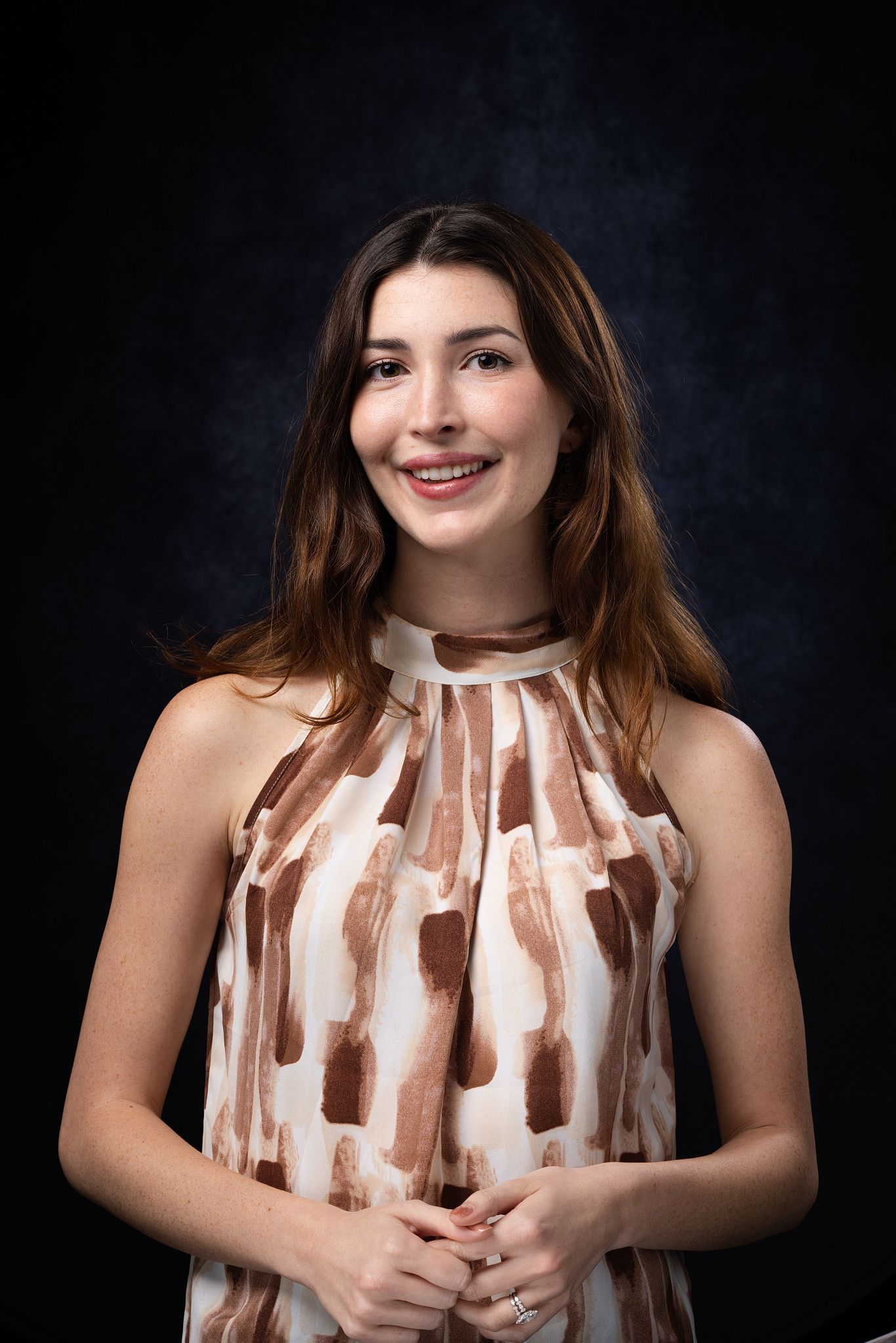 Smiling young woman with long brown hair wearing a white and brown patterned sleeveless dress against a dark background.