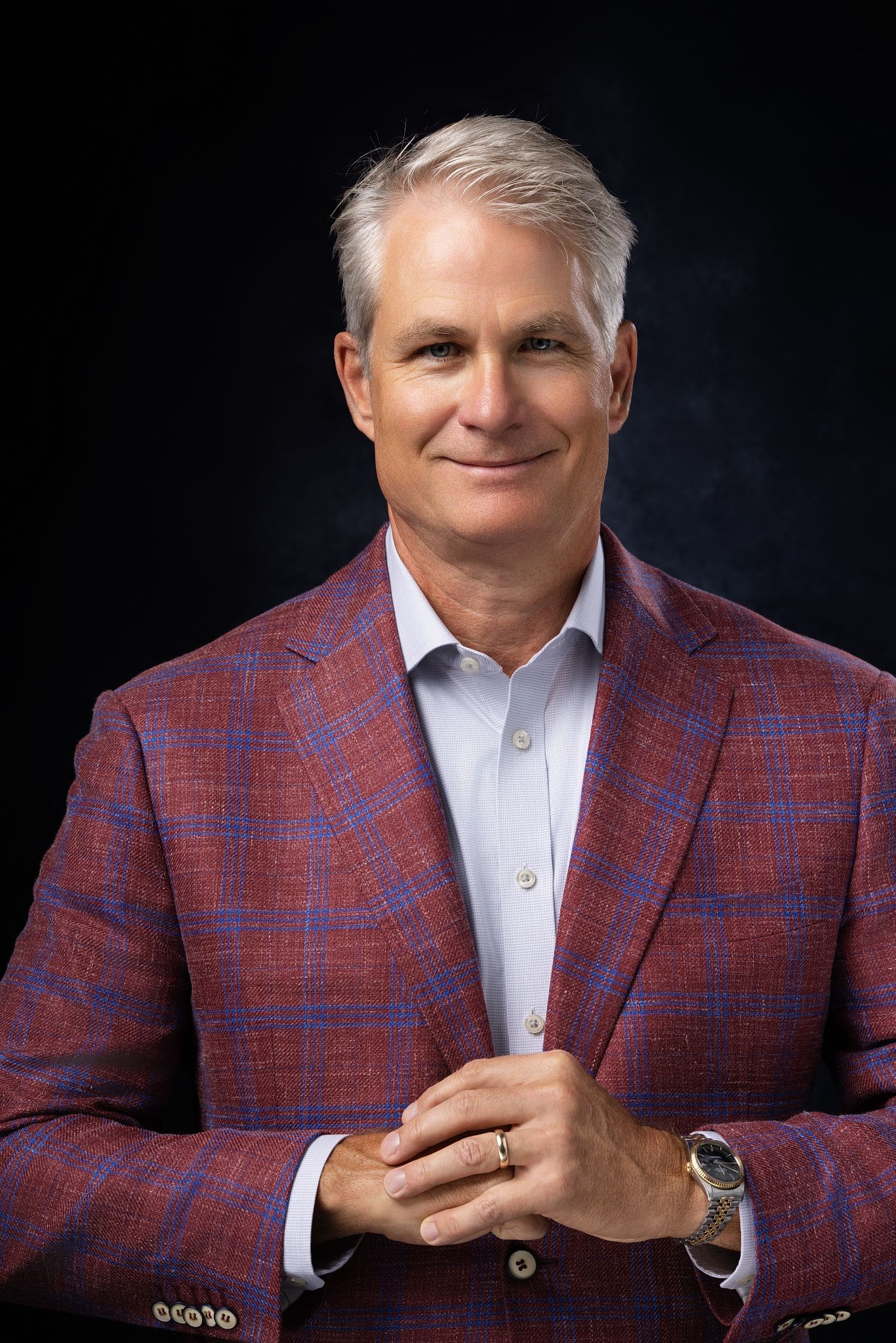 Professional man in red plaid blazer smiling confidently against dark background, wearing a watch and ring.