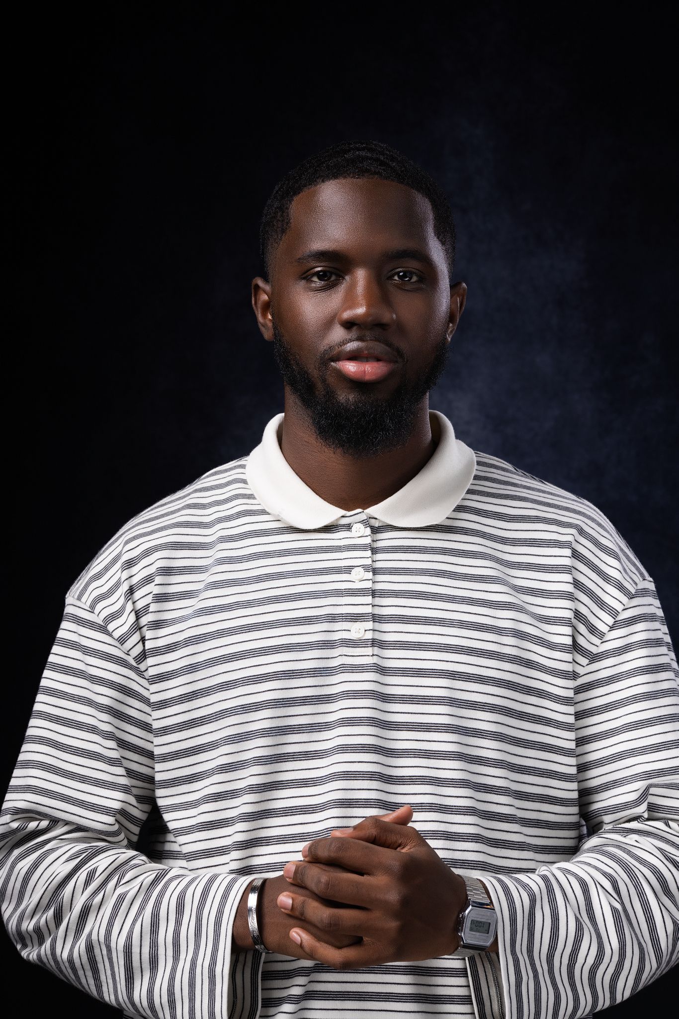 Young Black man with beard wearing striped polo shirt, hands clasped, posing against dark studio background.