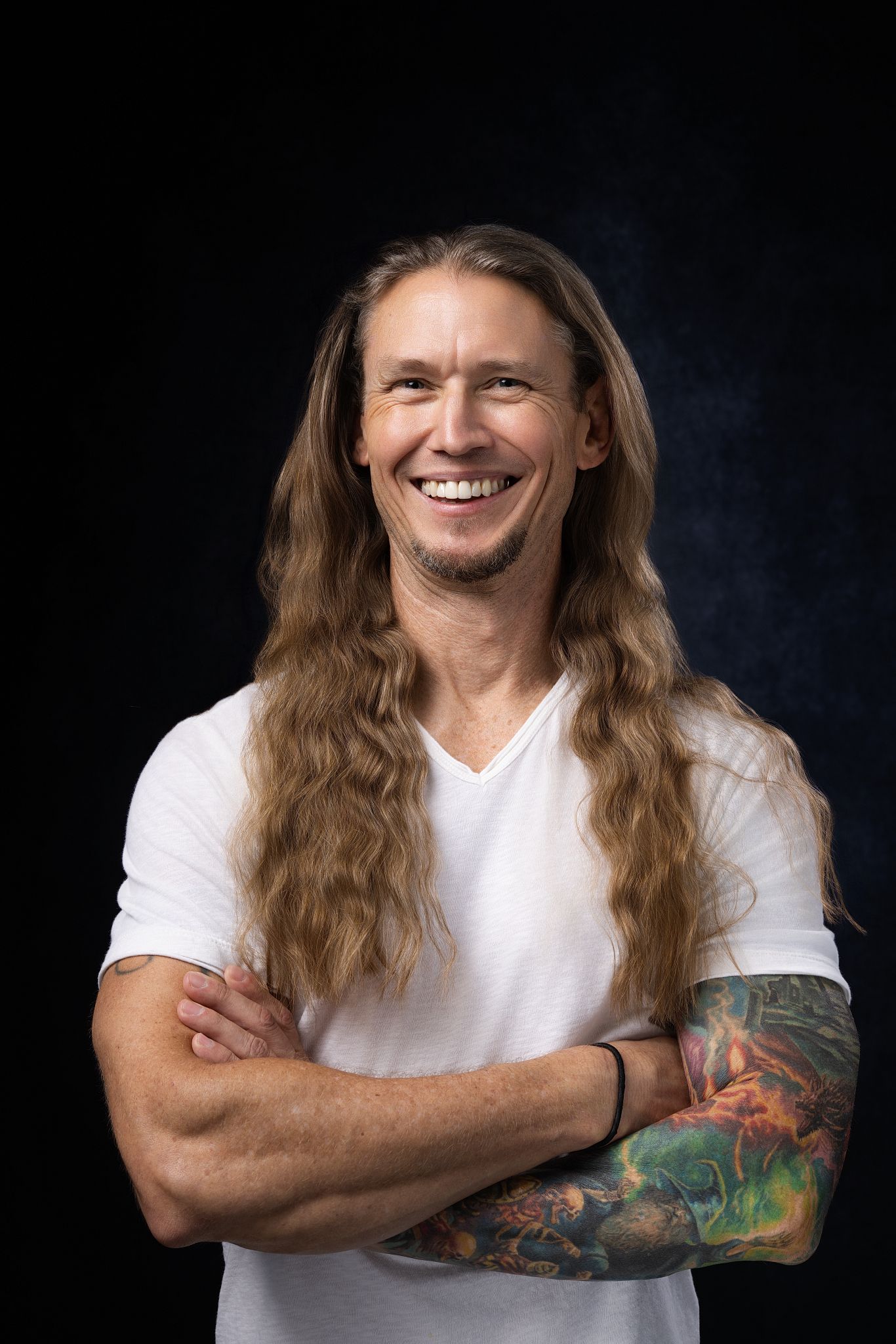 Smiling man with long brown hair, tattoo sleeve, and white V-neck t-shirt posing with arms crossed on dark background.