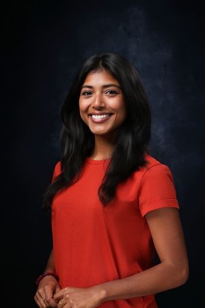 Smiling young woman with long dark hair wearing a red top, posed against a dark studio background.