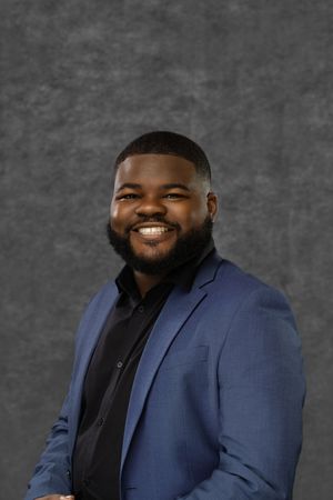 Smiling young man with beard wearing a blue blazer and black shirt posing against a gray background.