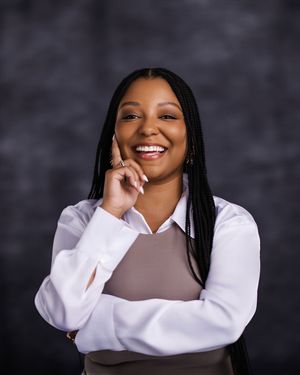 A Black woman with long braids smiles brightly while posing in a white shirt and brown vest.