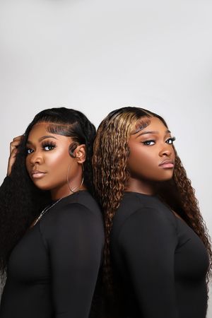 Two Black women with long curly hair in black outfits pose back-to-back against white background.