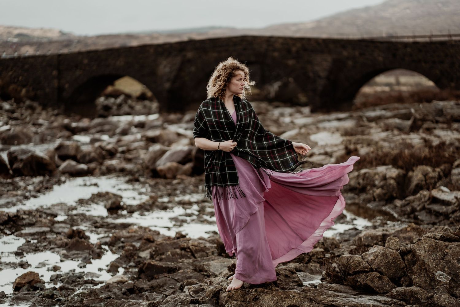 Figure in a black plaid top and flowing mauve skirt stands gracefully among rocky coastal terrain at dusk.