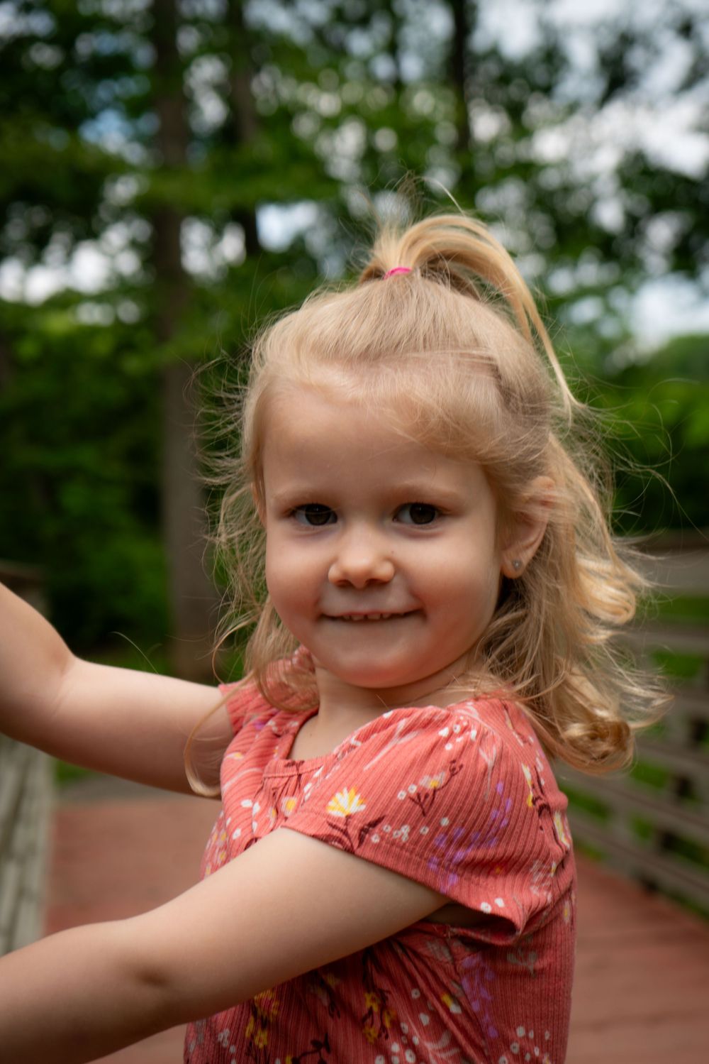 A toddler in a coral-colored dress smiles while standing on a wooden bridge outdoors with trees in the background.