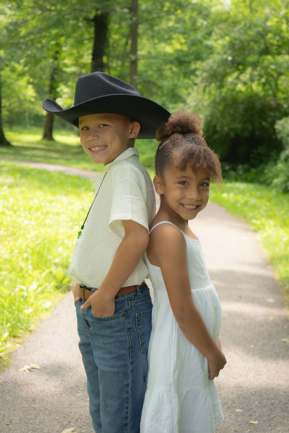 Two children standing back to back on a sunny path in a park, one wearing a cowboy hat and jeans, other in white dress.