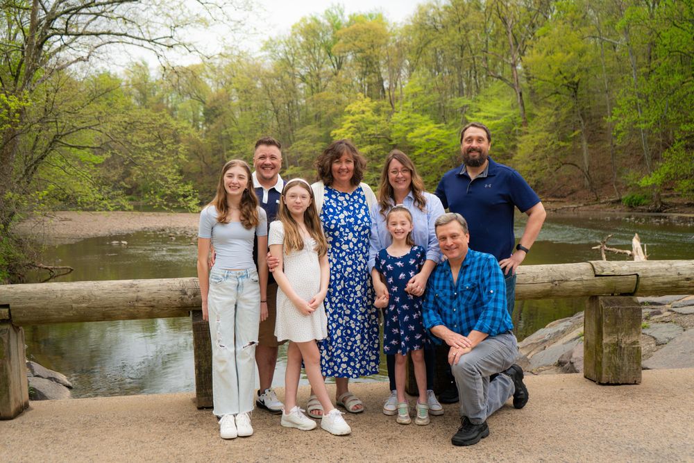 Family group poses together in a natural park setting with trees and a lake in the background on a sunny day.