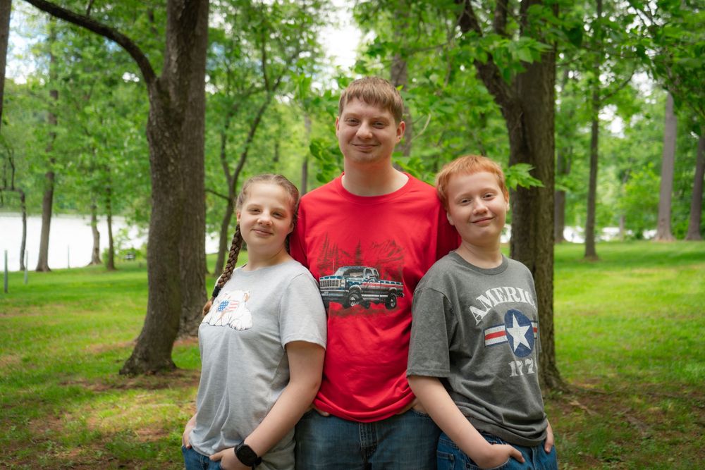 Three people in casual t-shirts stand together smiling in a green park with trees and a lake in the background.