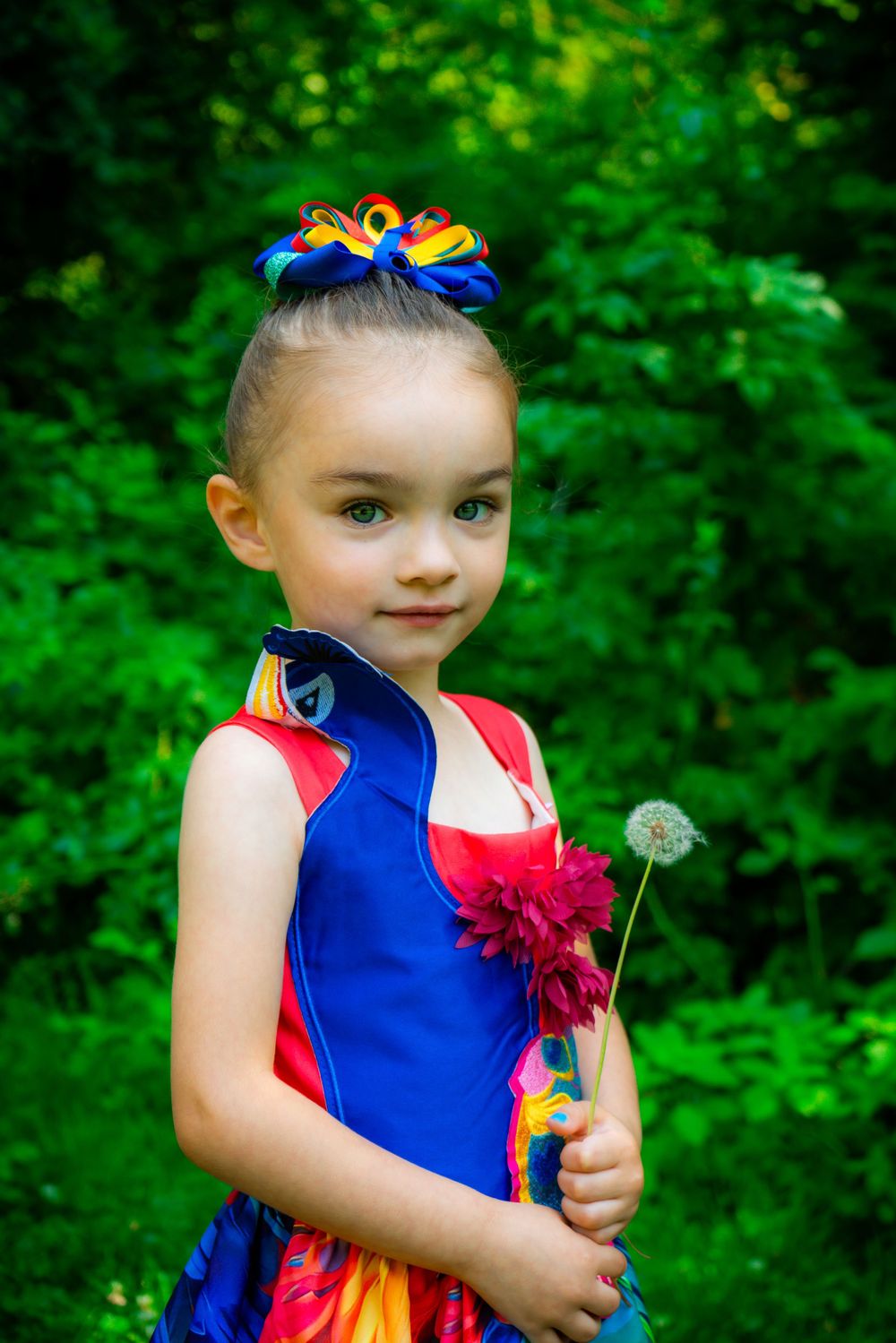 Young performer in colorful dress holds dandelion while posing against lush green background.