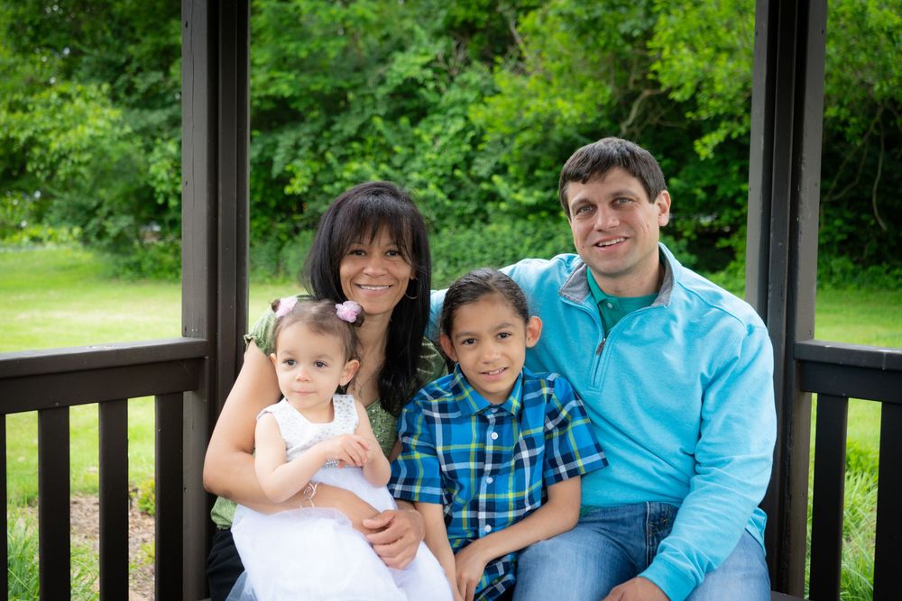 A smiling family sits together on a wooden deck railing surrounded by lush green trees on a sunny day.