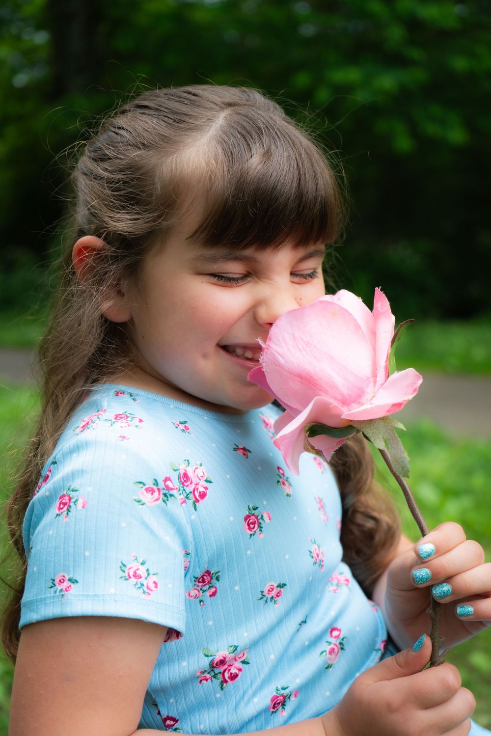 A young smiling figure wearing a blue floral shirt smells a large pink peony flower in an outdoor garden setting.