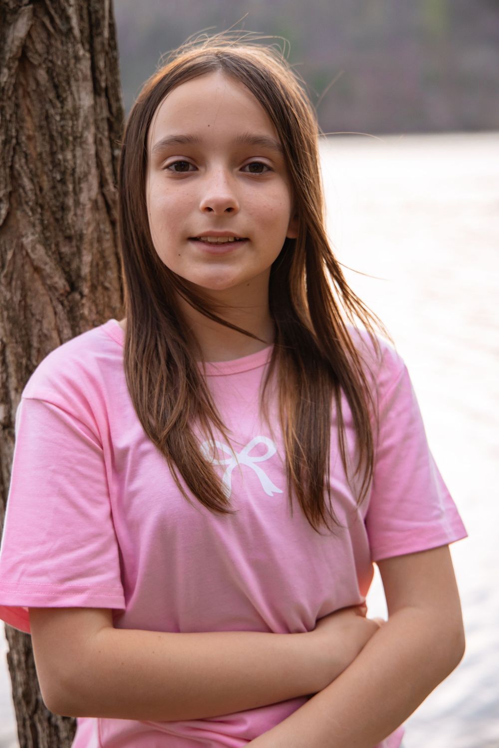 Young person wearing a pink t-shirt stands next to a tree trunk outdoors on a bright day.