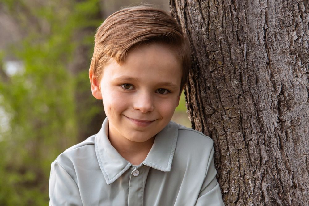 A young person wearing a light gray collared shirt leans against a large tree trunk in an outdoor setting.