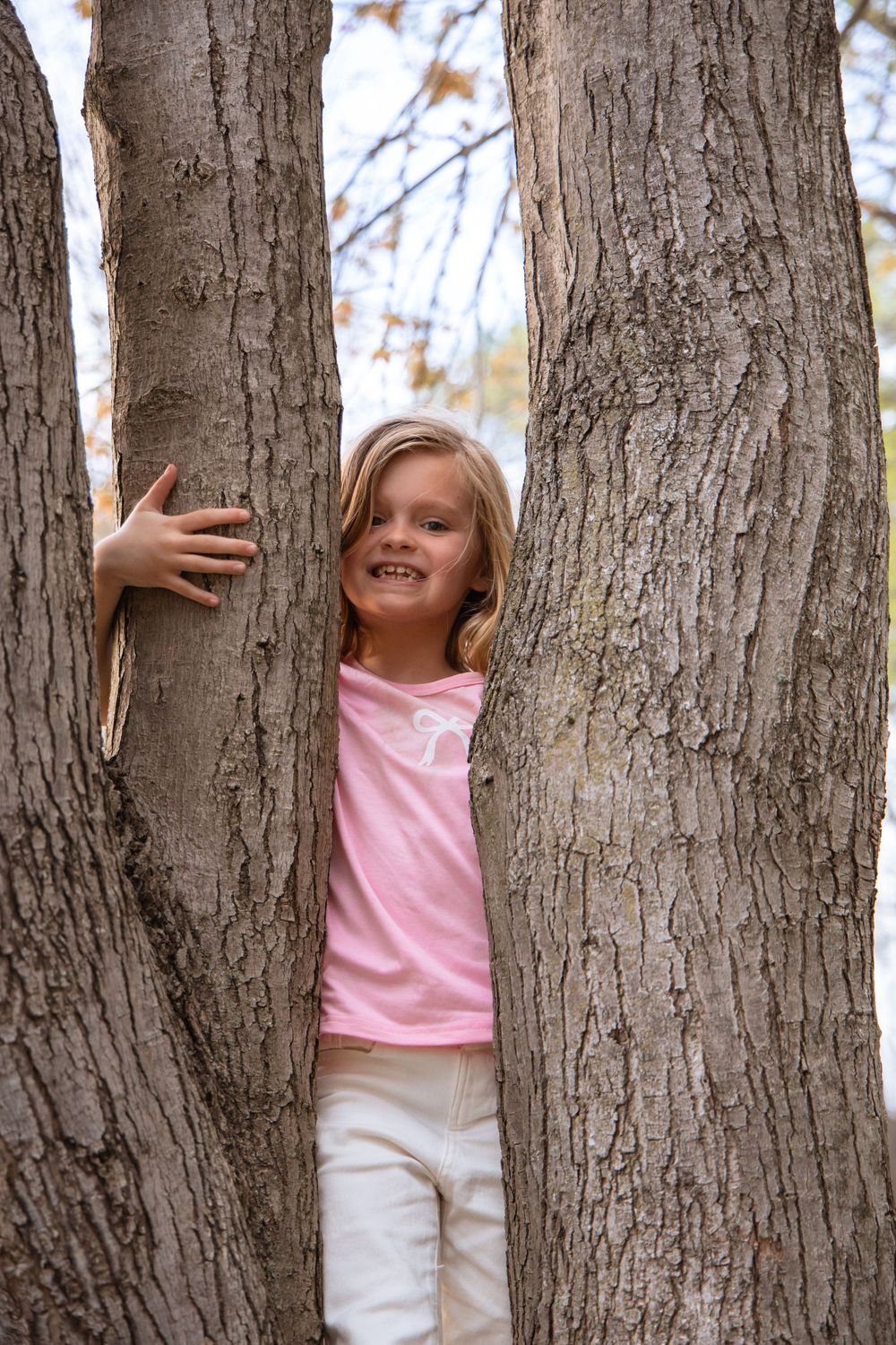 A child in a pink shirt and white pants plays between large tree trunks in a wooded area.