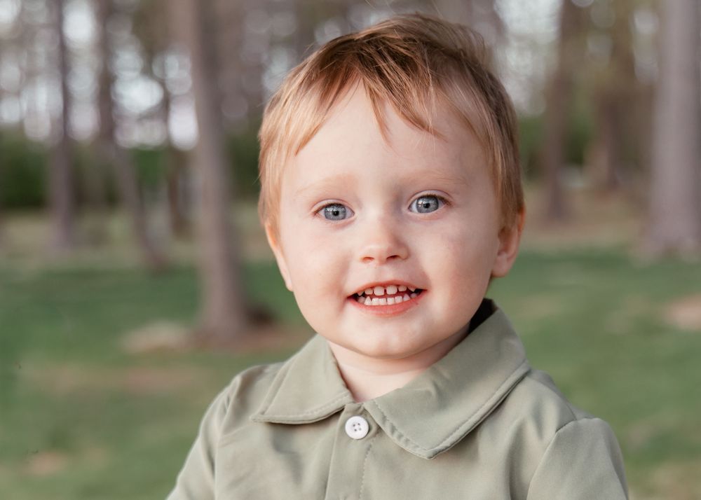 A smiling child wearing a sage green jacket poses outdoors against a blurred natural backdrop.