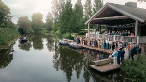 A scenic lakeside wedding ceremony setup with guests seated at a covered outdoor venue at sunset.