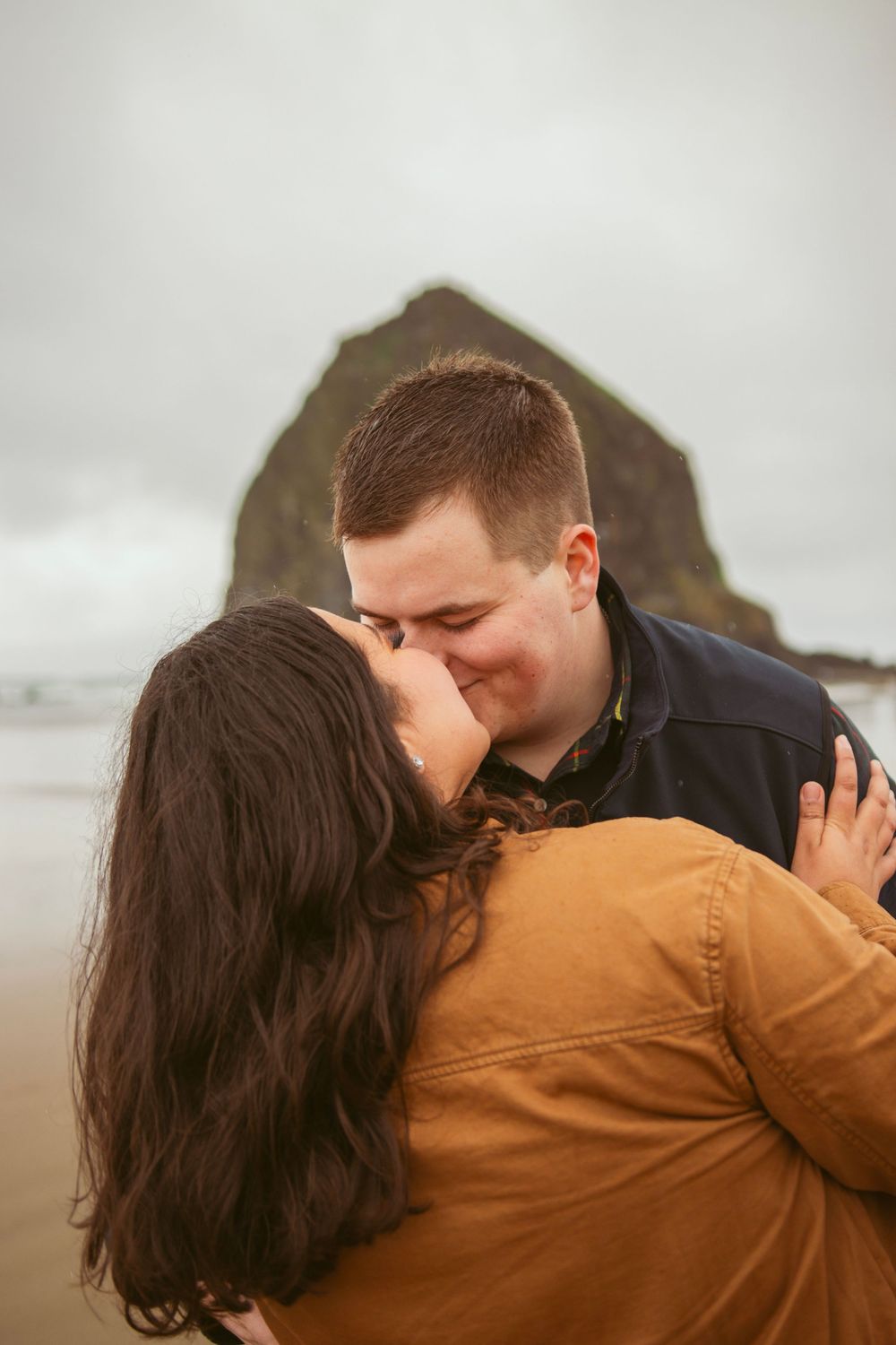 Couple sharing an intimate moment and embracing on a misty beach with a large rock formation in the background.