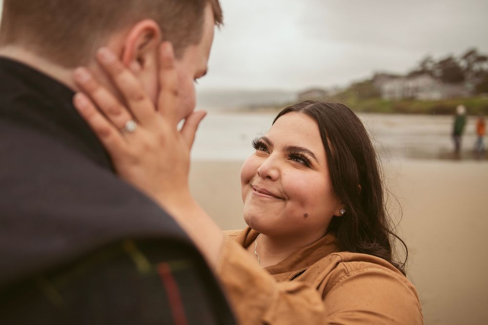 Two people share an intimate emotional moment on a cloudy beach with someone in the background.
