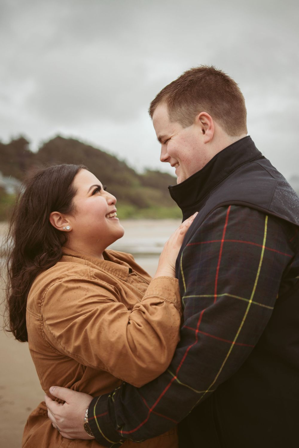 A couple embraces lovingly on a cloudy beach day, wearing fall jackets and sharing a tender moment together.