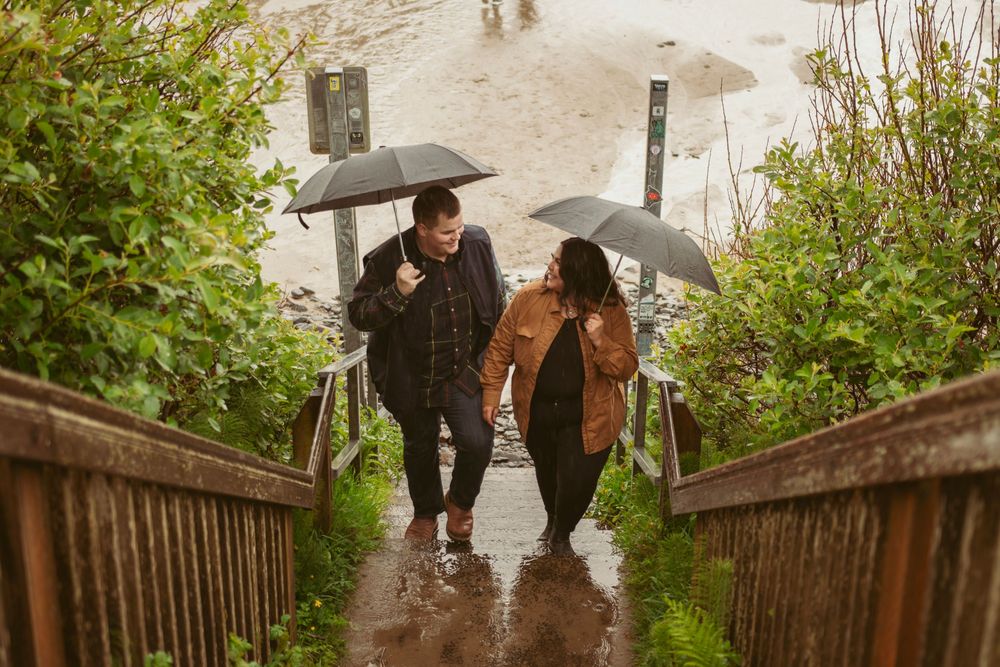 People with umbrellas climb wooden stairs on a rainy day at a beach while enjoying each other's company.
