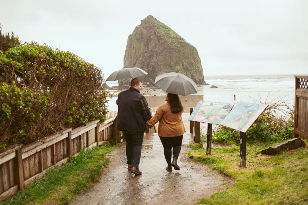 Two people with umbrellas walk on a rainy path overlooking Haystack Rock at Cannon Beach, Oregon on a stormy day.