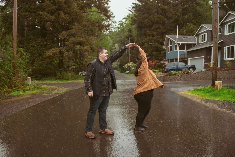 Two people dance together holding hands on a wet suburban road during a rainy day with evergreen trees in background.