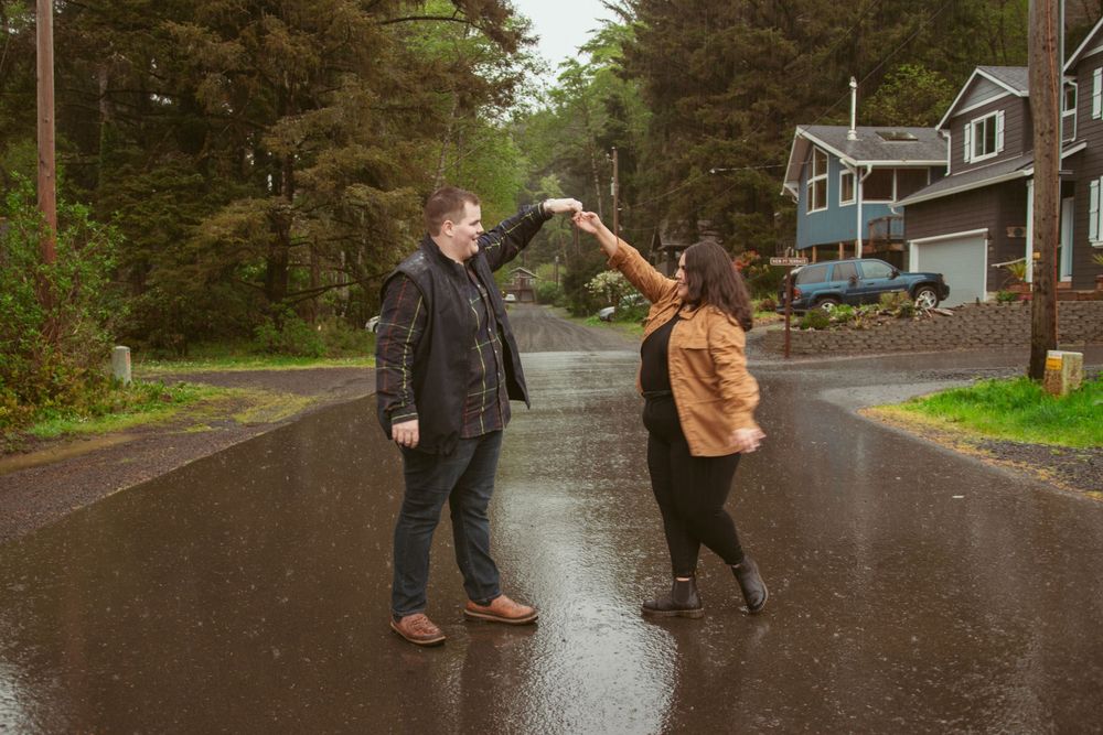 Two people playfully dance together on a wet rural road in front of a grey house on a rainy day.