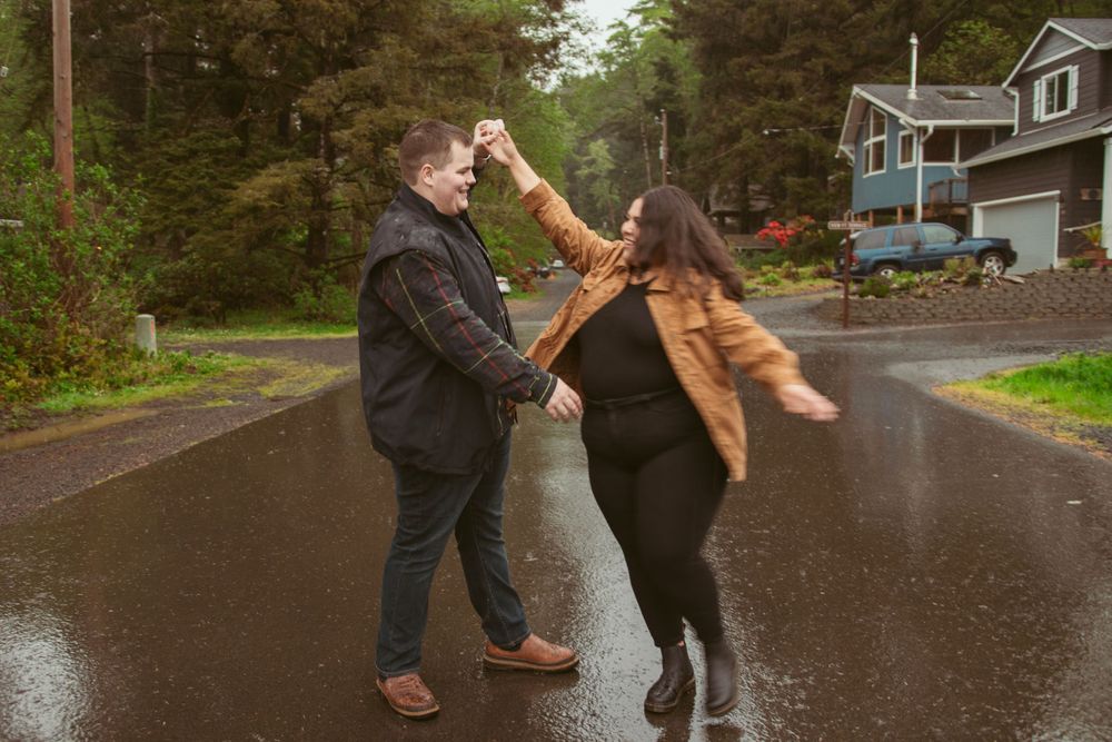 Two people dance together playfully on a wet residential street surrounded by trees and houses.