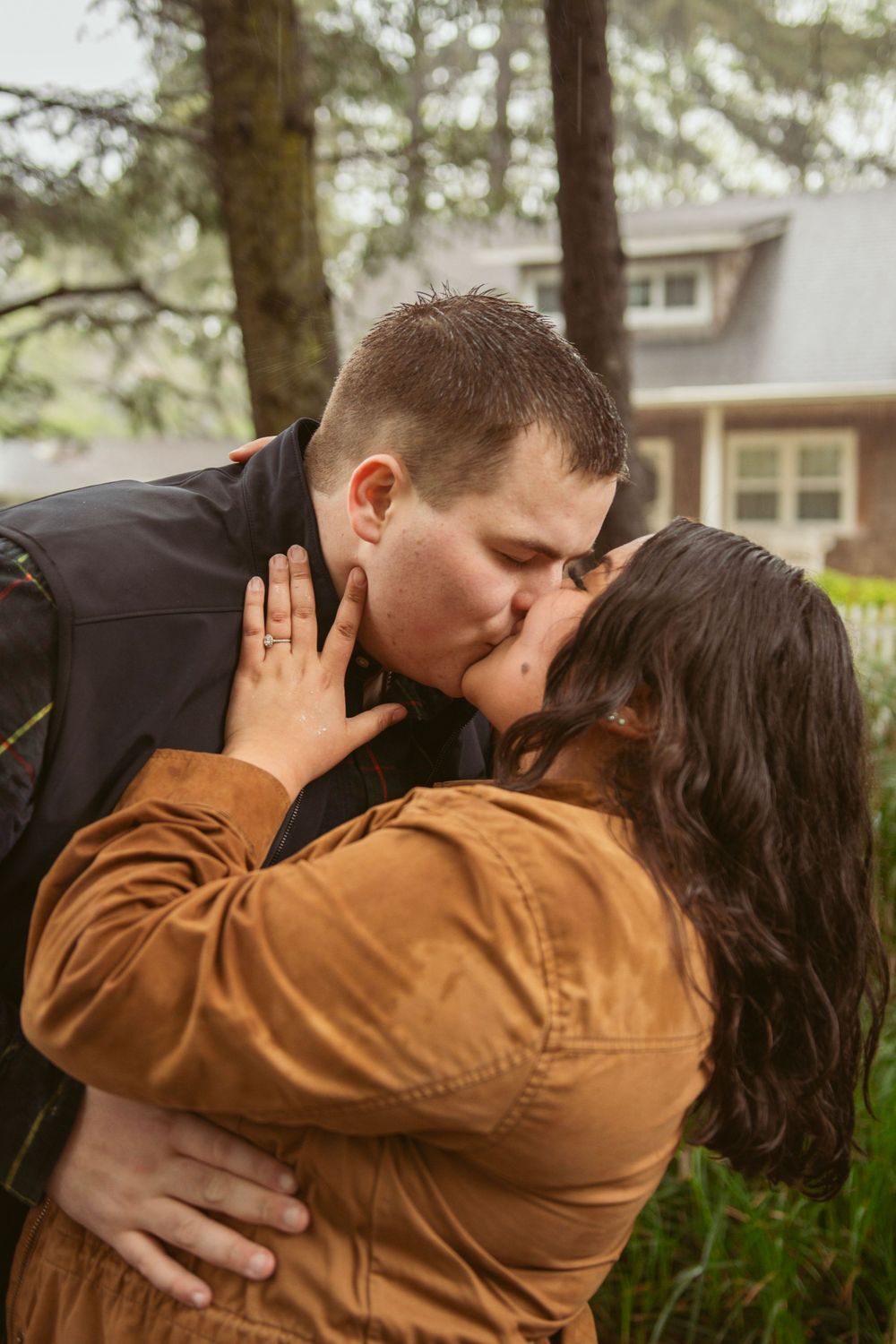 A couple shares a romantic kiss outdoors with a house in the background among trees on a cloudy day.
