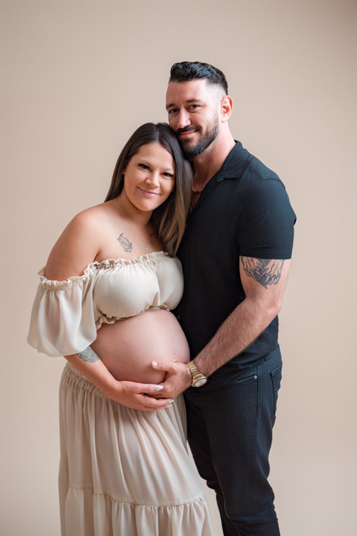 An intimate maternity photo session features a couple posing in white and black against a neutral backdrop.
