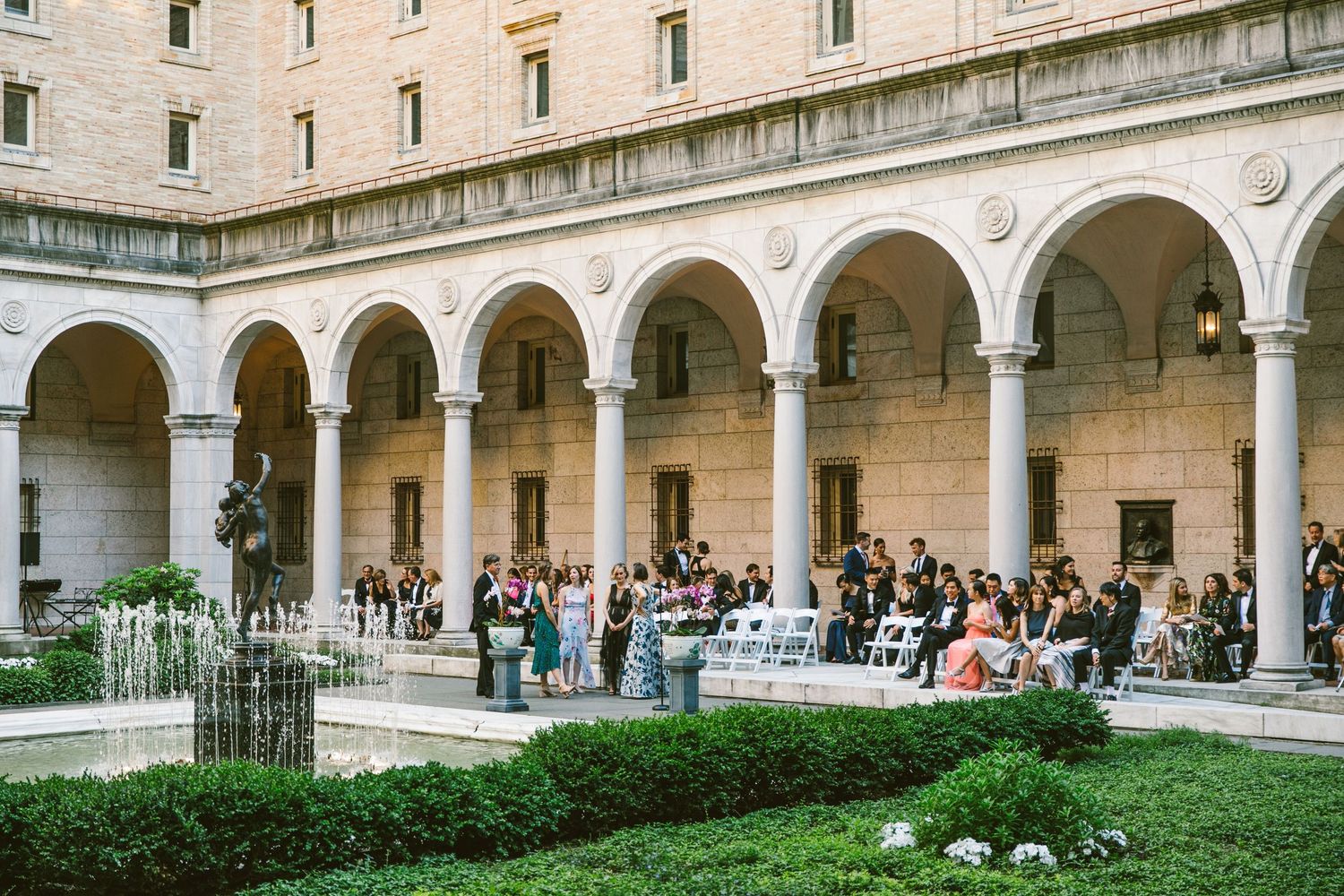 People gather in an elegant courtyard with arched stone colonnade and manicured gardens at The Boston Public Library.