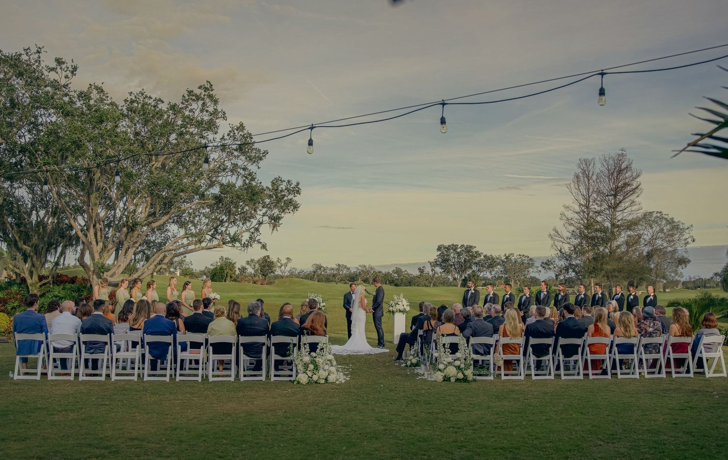 Panoramic view of an outdoor wedding ceremony setup with rows of white chairs and guests on a grassy hillside at sunset.