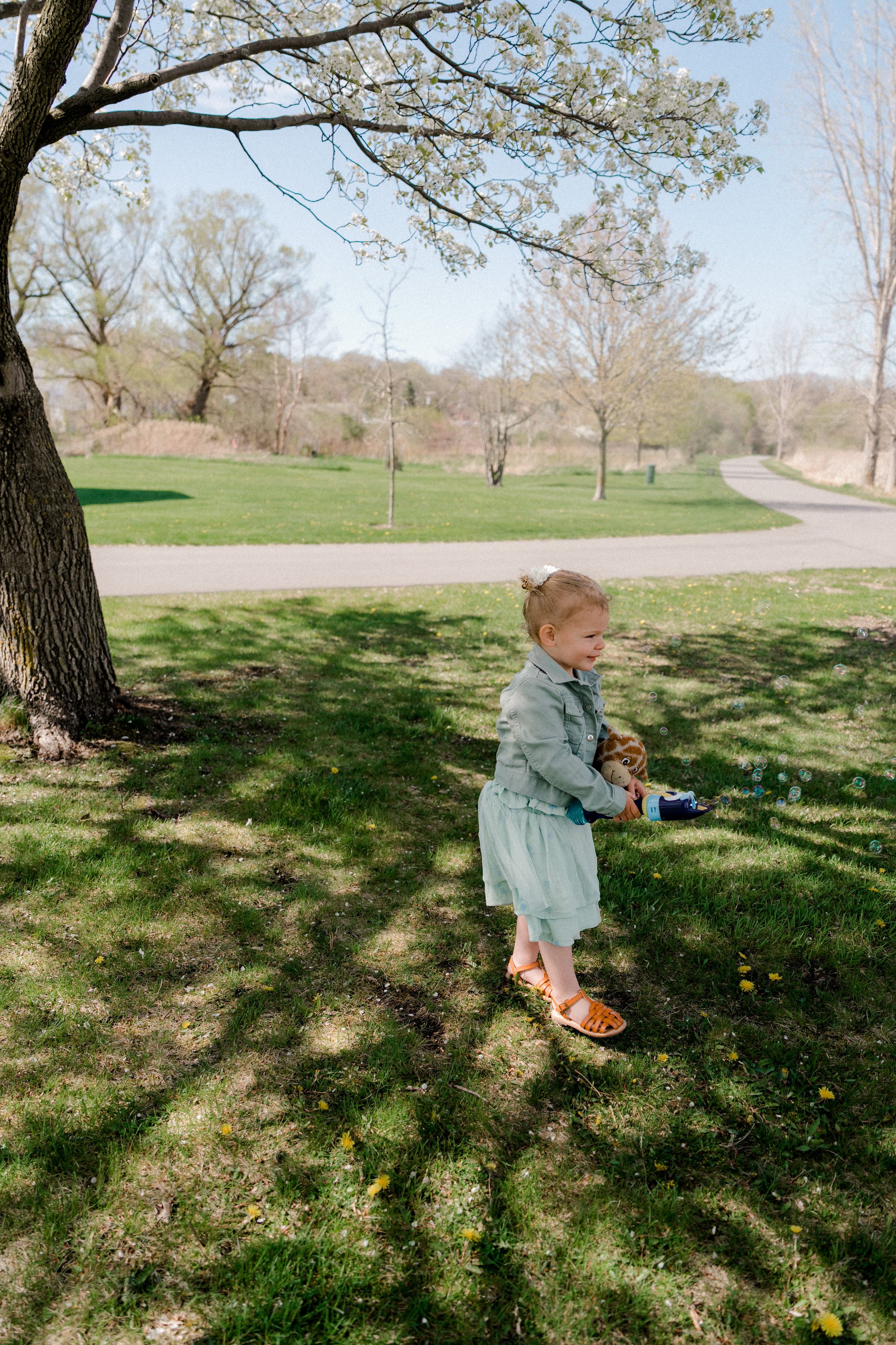 A small child in a denim dress plays in the dappled shade beneath a blossoming spring tree.